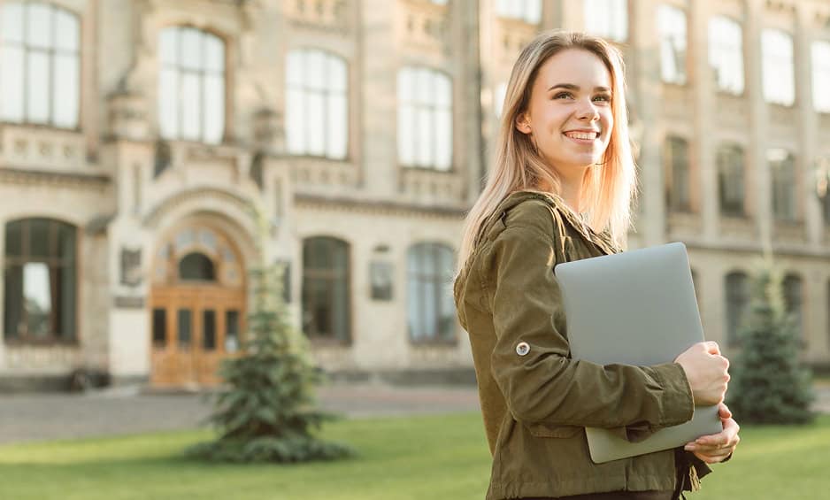 Happy female student holding laptop on university campus in outdoor setting.
